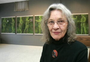 photograph of Bernice Vincent. Her head and shoulders take up the right half of the frame. She has shoulder length, greying, wavy hair and glasses. She's wearing a black sweater with a brooch-pin that is a small painting. Behind her and taking up the left half of the frame are a few panels of her work "14 Women."